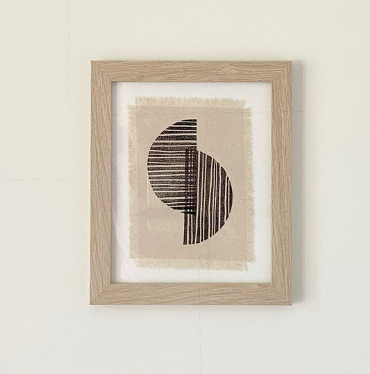 Block printed text in a frame.  Black half circles overlapping on off white frayed canvas.  Natural colored frame.
